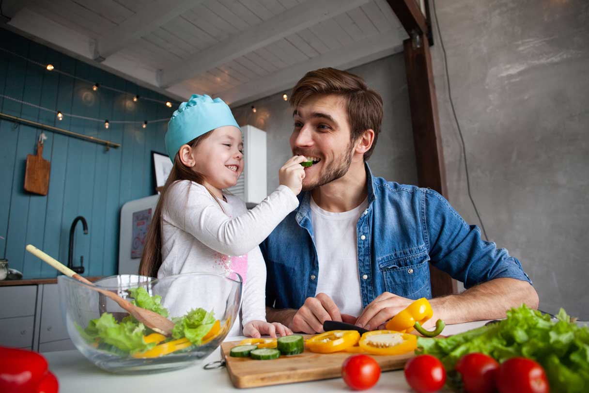 Man preparing a salad with his young daughter as she feeds him a piece of food