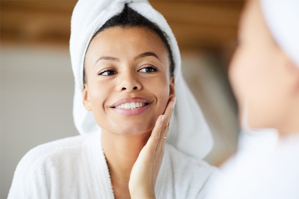 Woman with white towel on her head, smiling as she touches her face and examines her reflection
