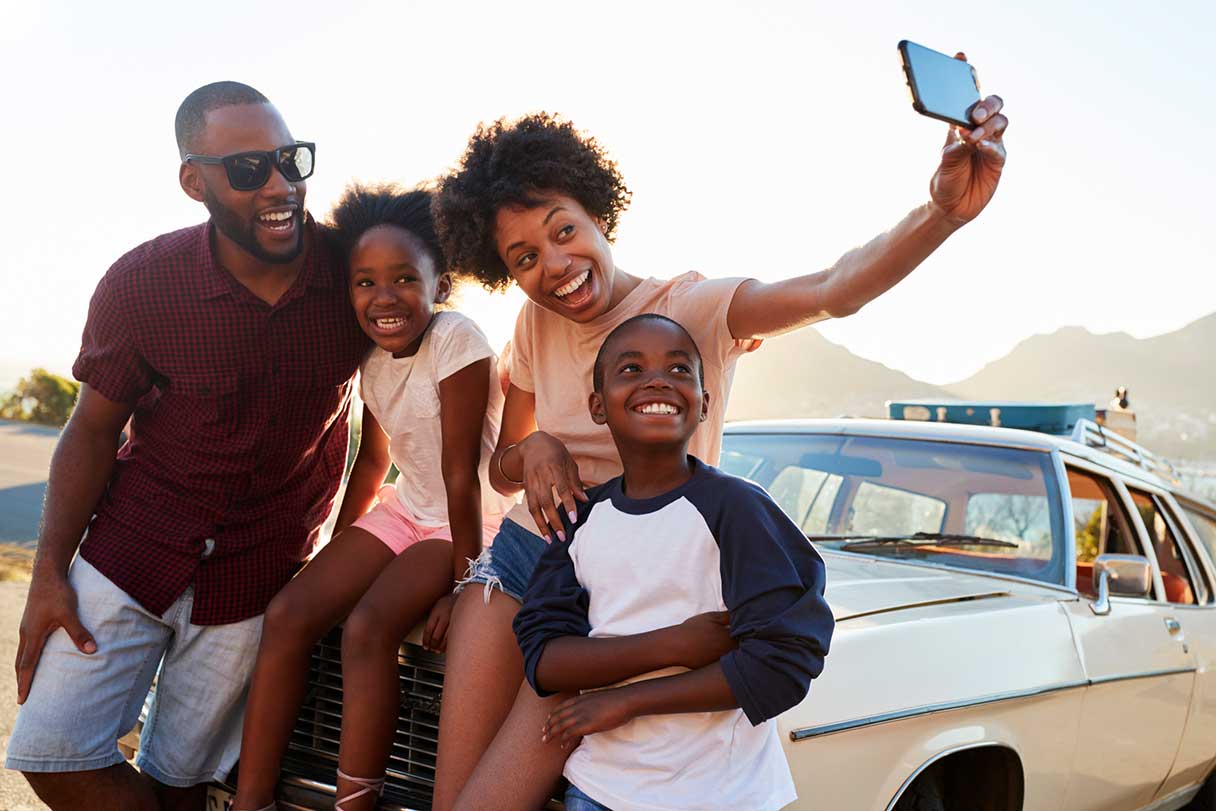 Family of four taking selfie while sitting on the hood of a car