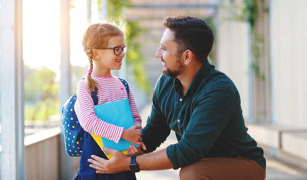 Man kneeling next to young girl wearing backpack and holding folders