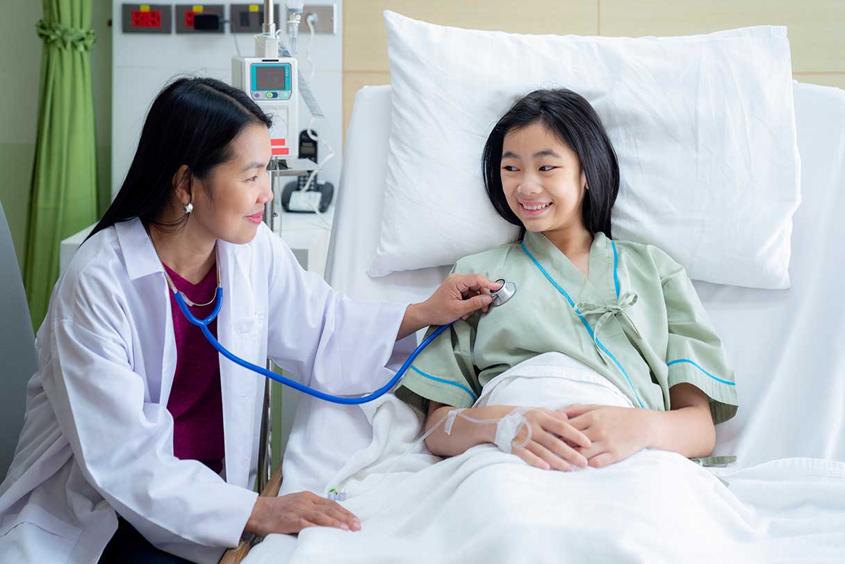 Young girl in hospital bed smiling at doctor inspecting her with a stethoscope