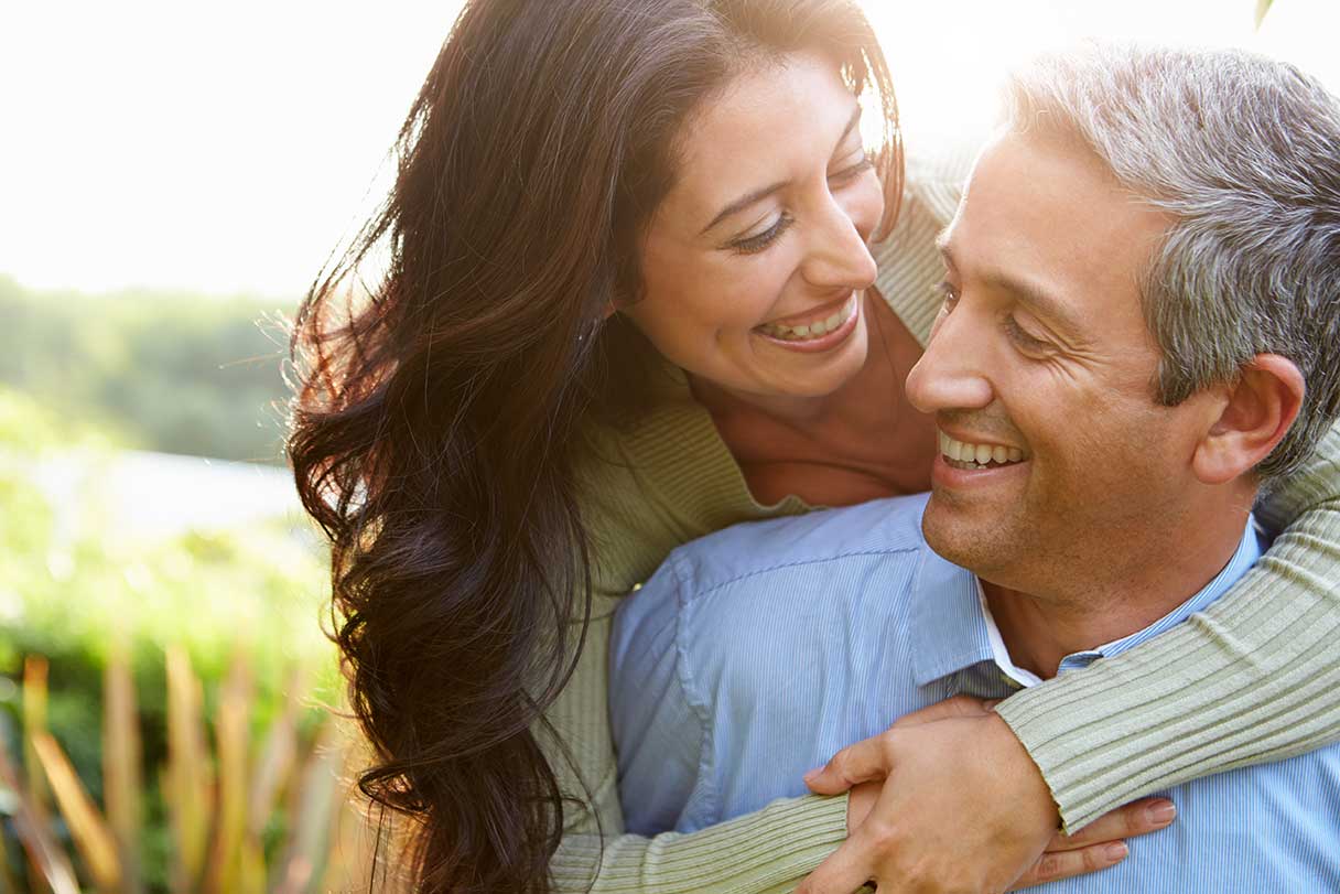 Woman with her arms around a man's shoulders, both smiling at each other