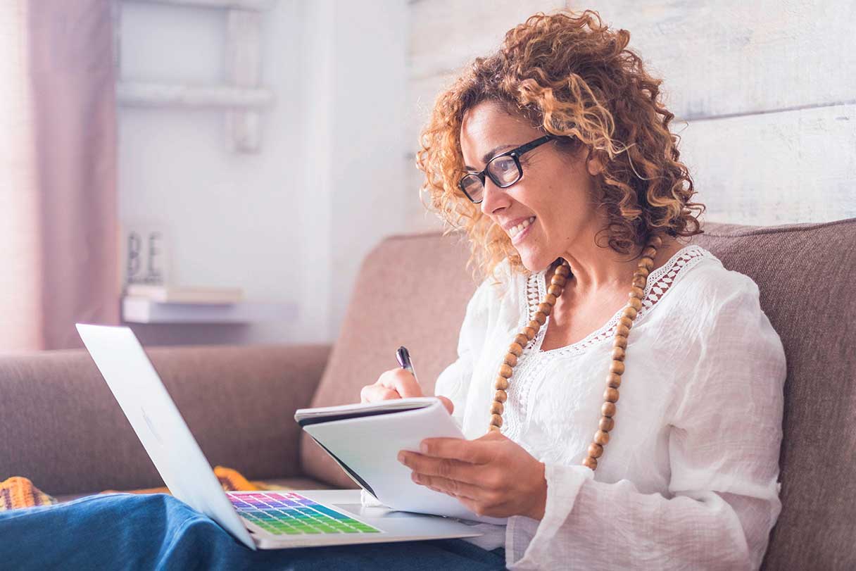 Woman in glasses holding notebook while she works on laptop