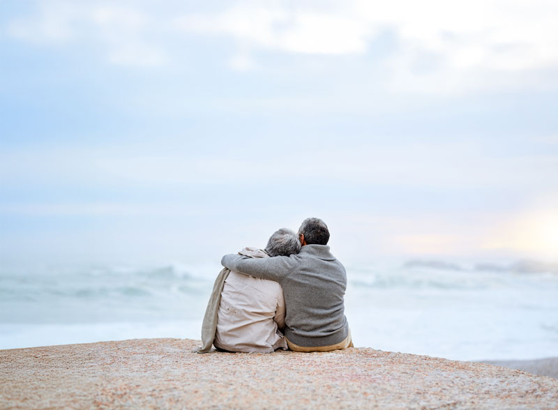 Man with his arms around a woman, both sitting on a beach