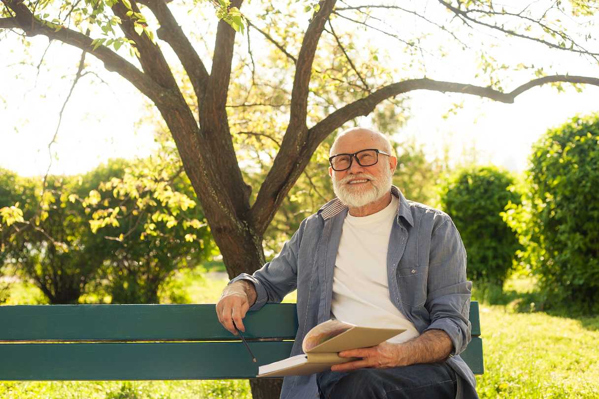 Man wearing glasses, holding a book and sitting on bench outside