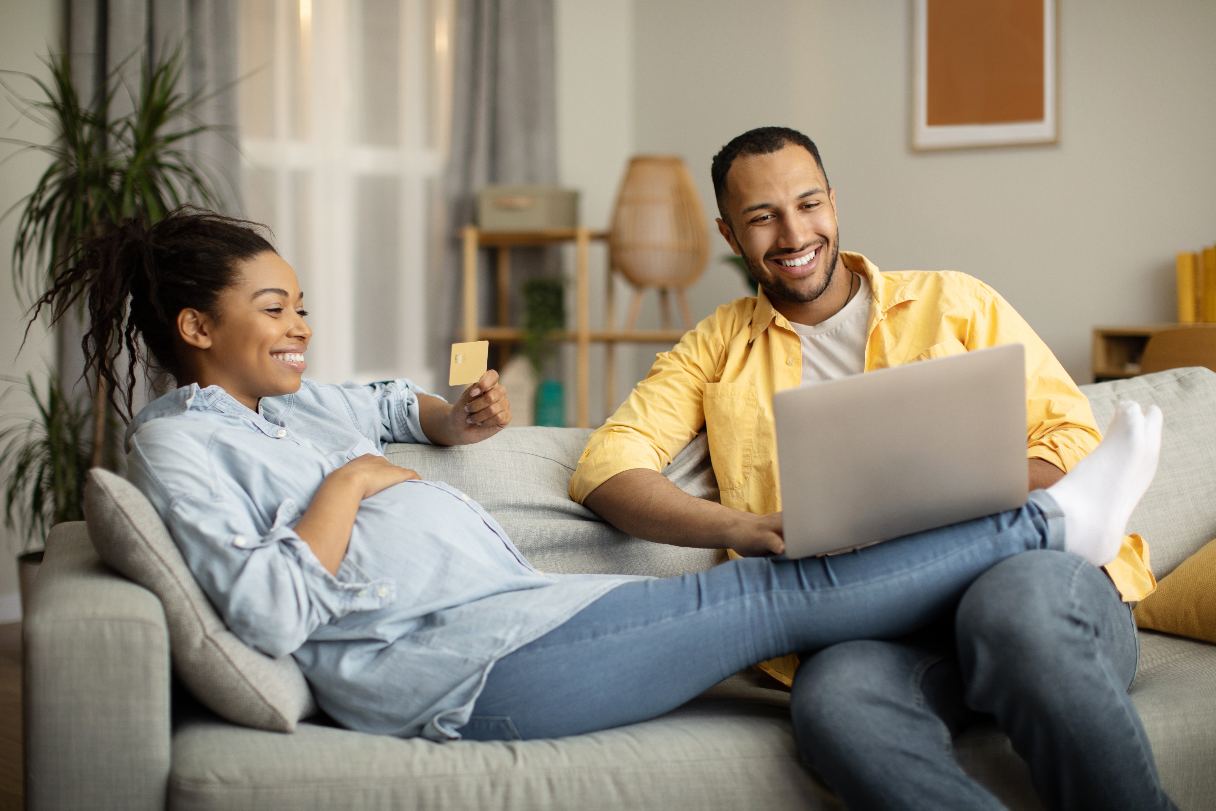 Pregnant woman holding credit card, sitting with man with laptop on sofa