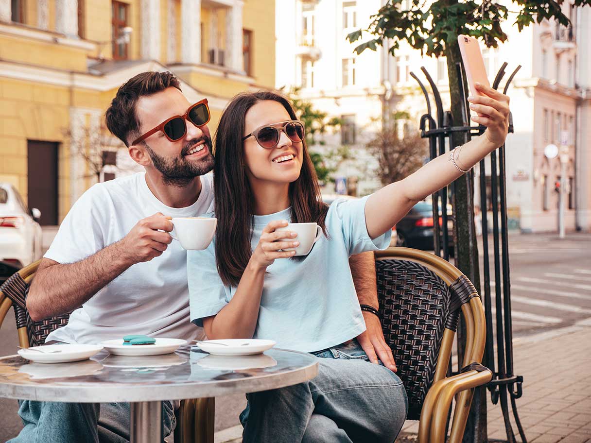 Man and woman in sunglasses sitting at outdoor cafe, taking selfie