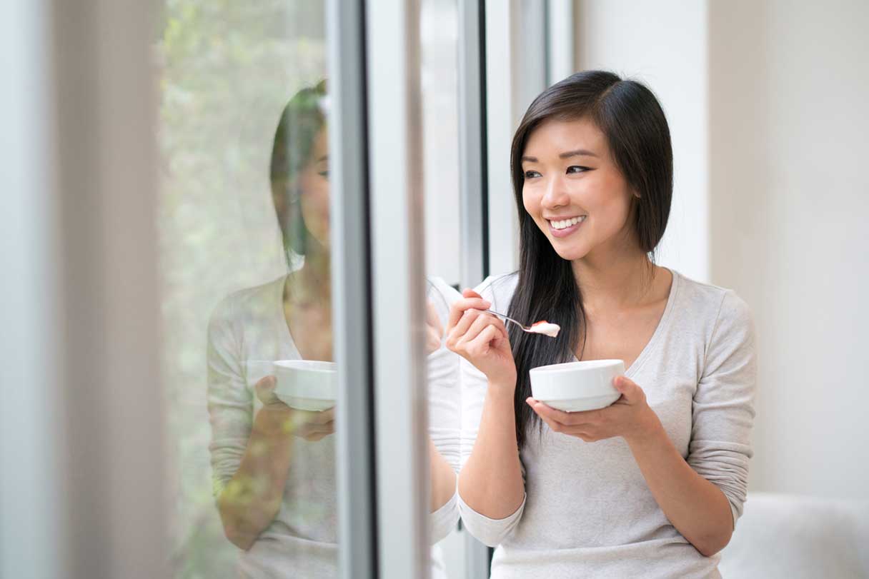 Woman smiling as she looks out window and eats out of a small bowl