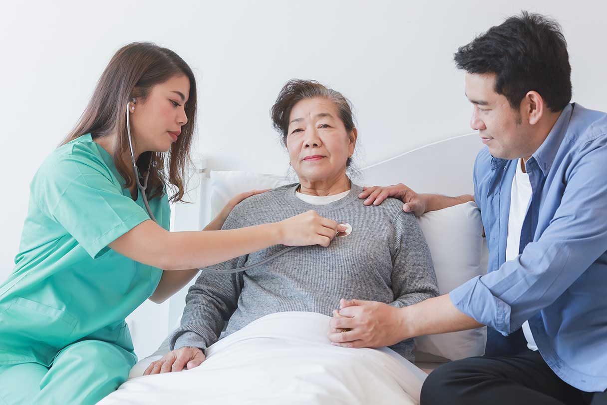 Nurse holding stethoscope to senior woman's chest, man sitting beside her hospital bed